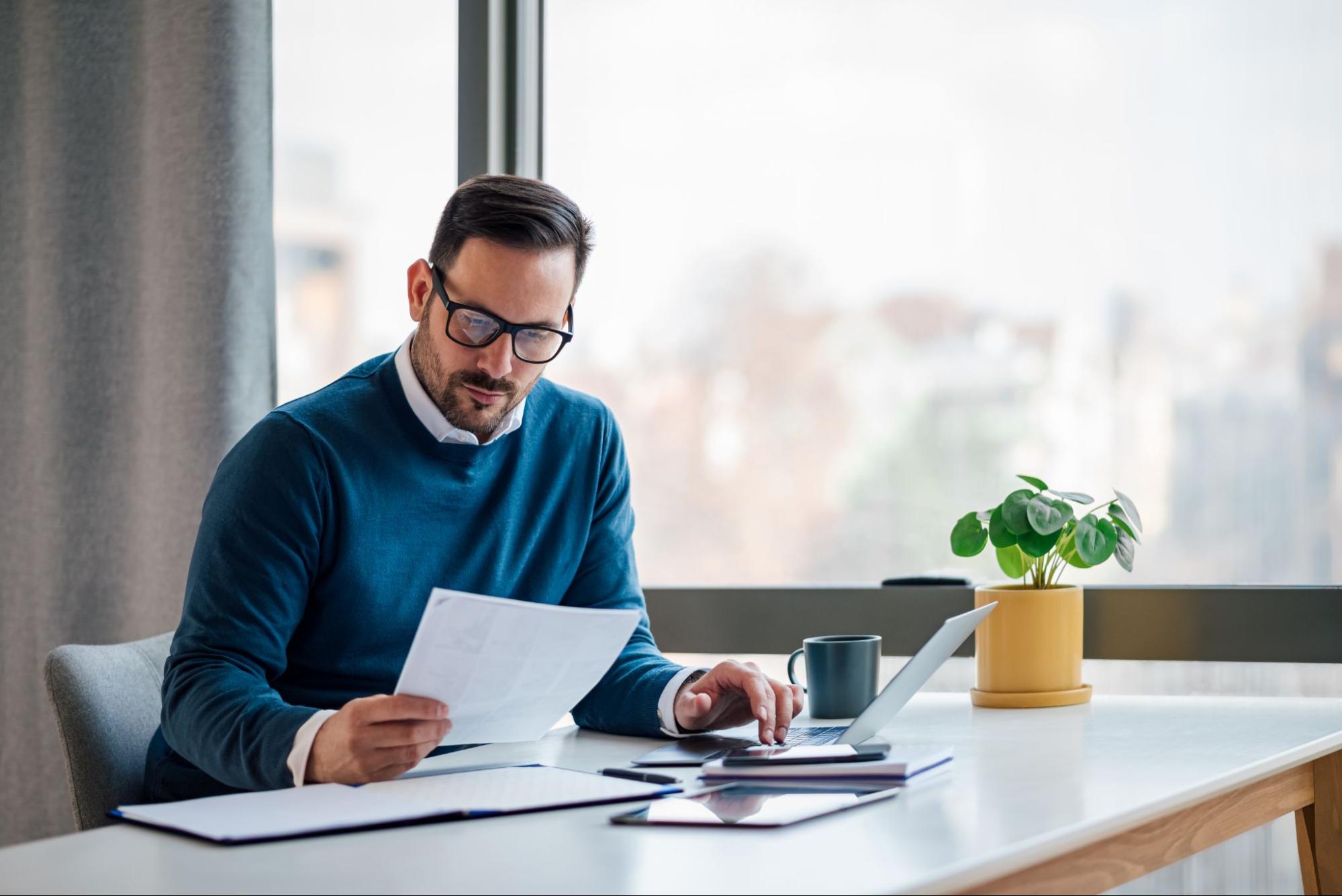Man in teal sweater looking at paper at desk